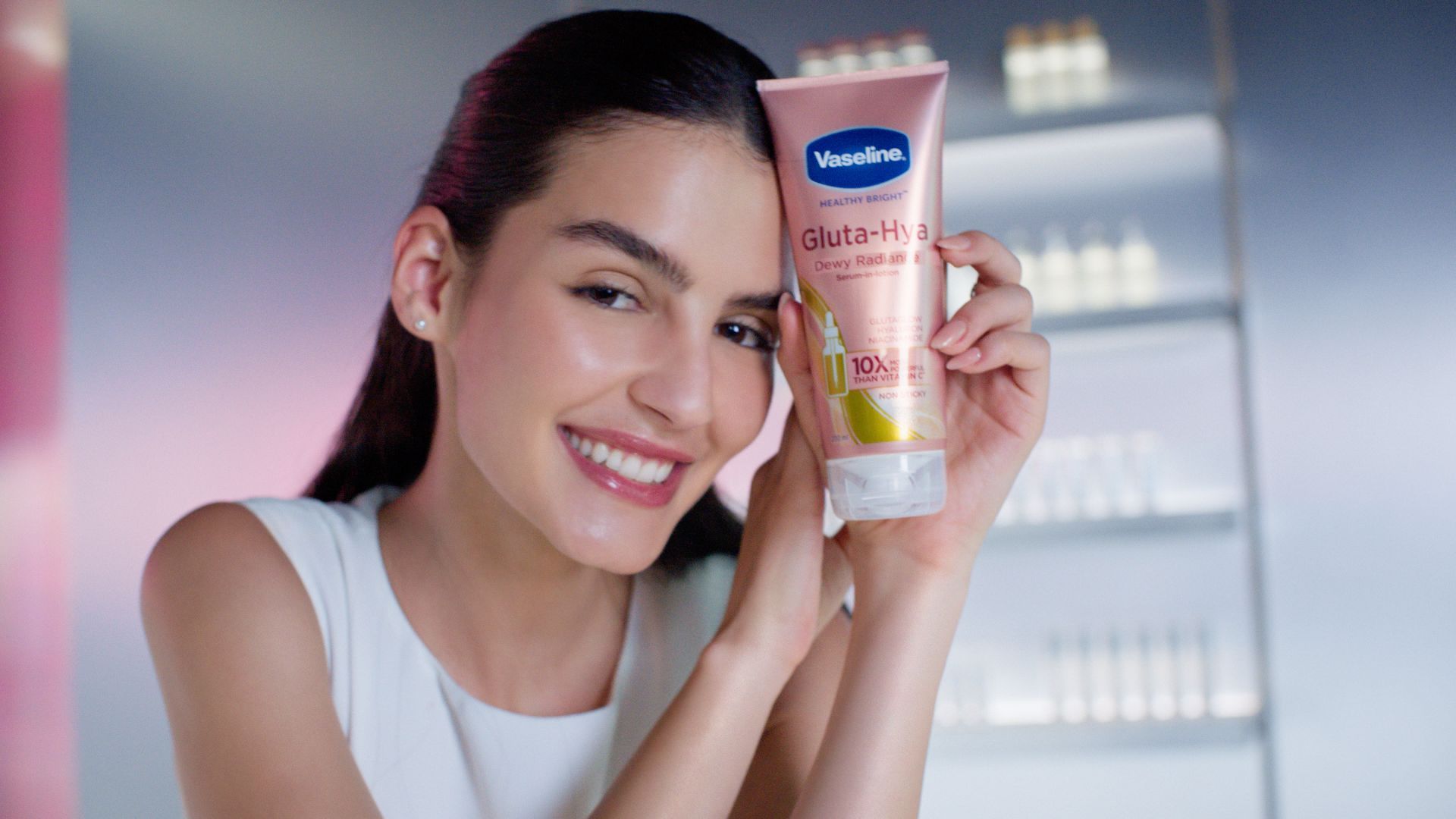 Uma mulher branca com cabelos escuros e pele brilhante, segurando um tubo de creme Gluta Hya Dewy Radiance da Vaseline. Embalagem premium cor-de-rosa.