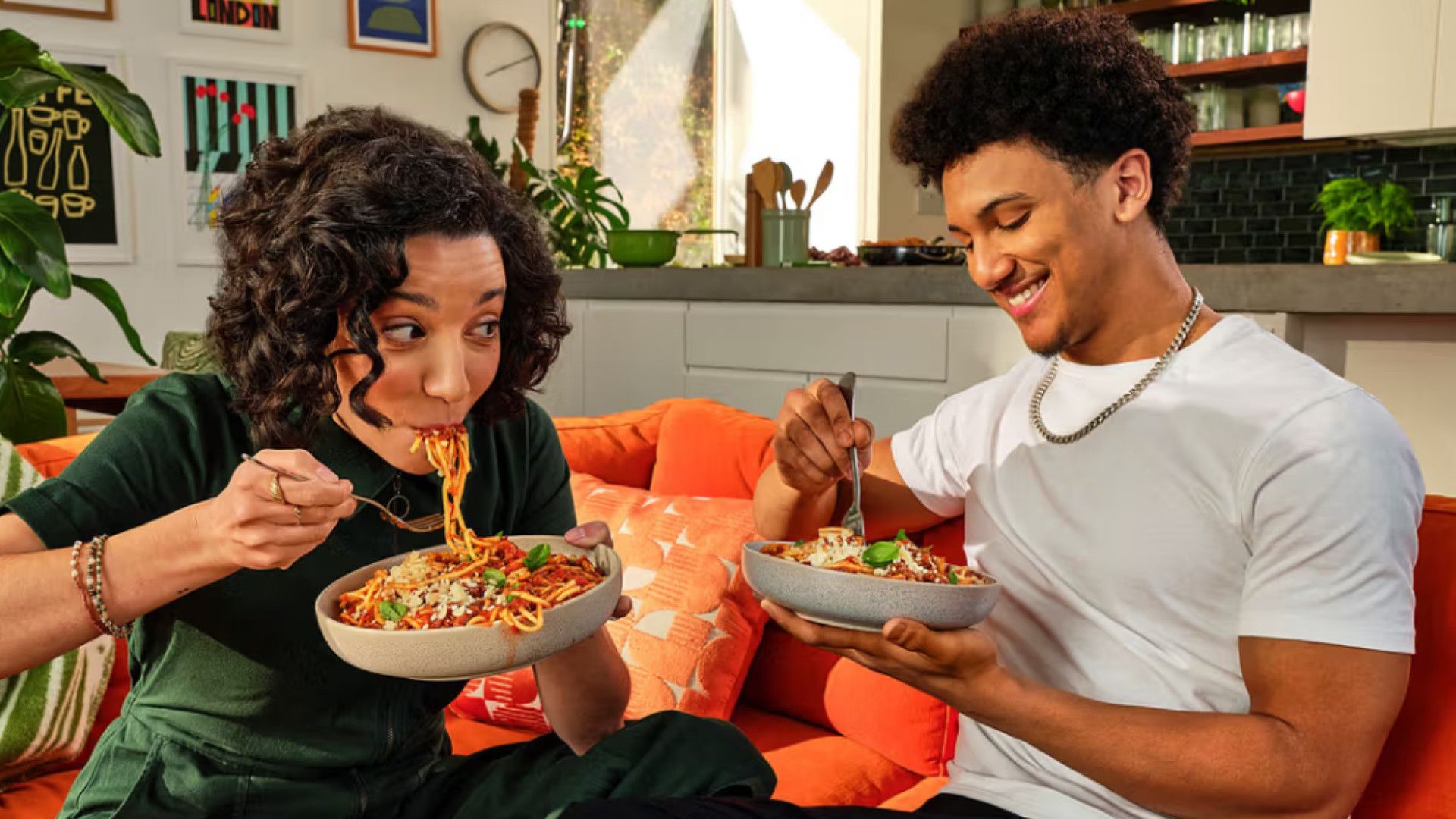 Duas pessoas sentadas em um sofá comendo tigelas de espaguete.