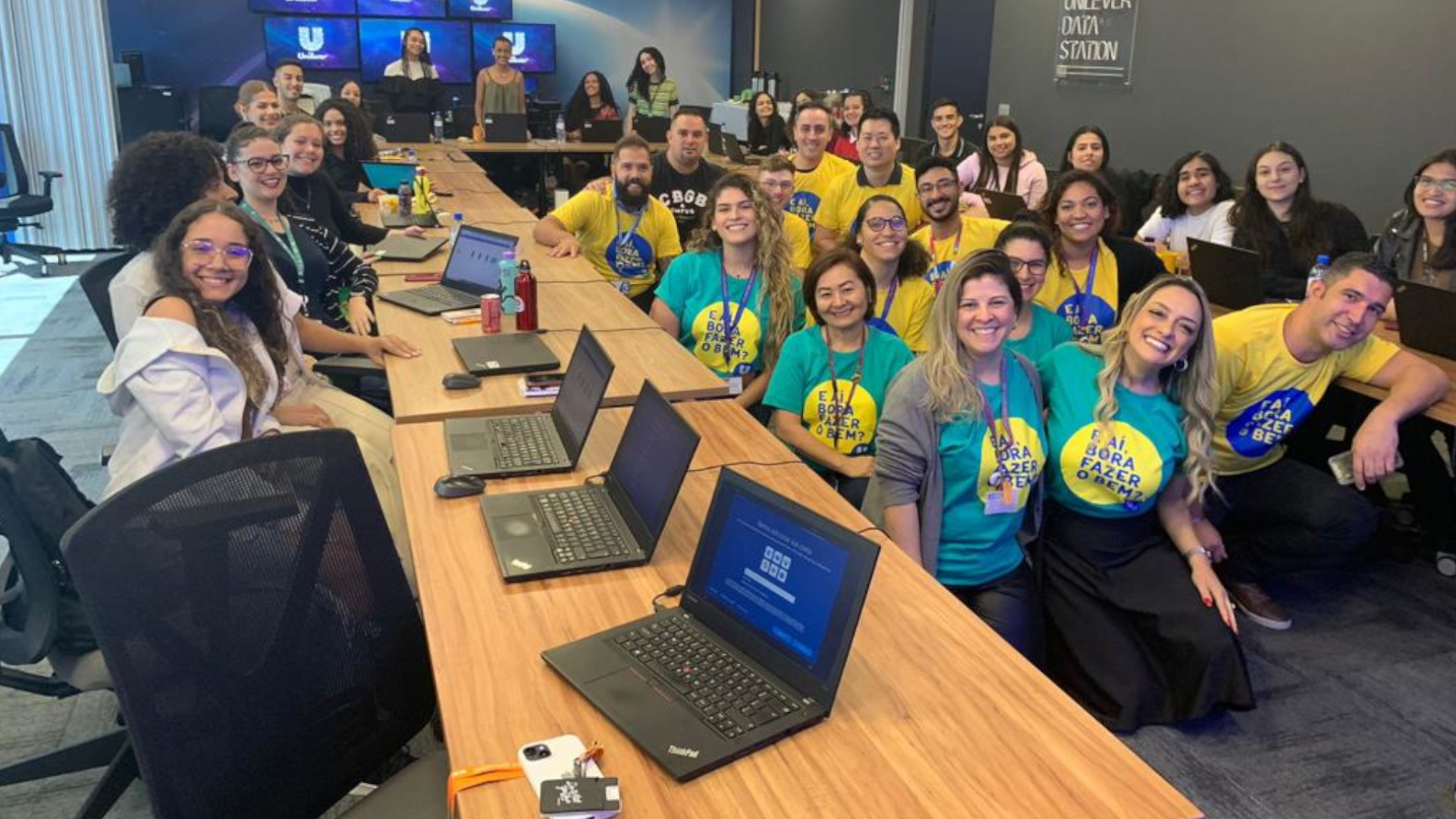 Jovens e funcionários da Unilever, com camisetas verdes e amarelas, sentados e alguns computadores em suas mesas.