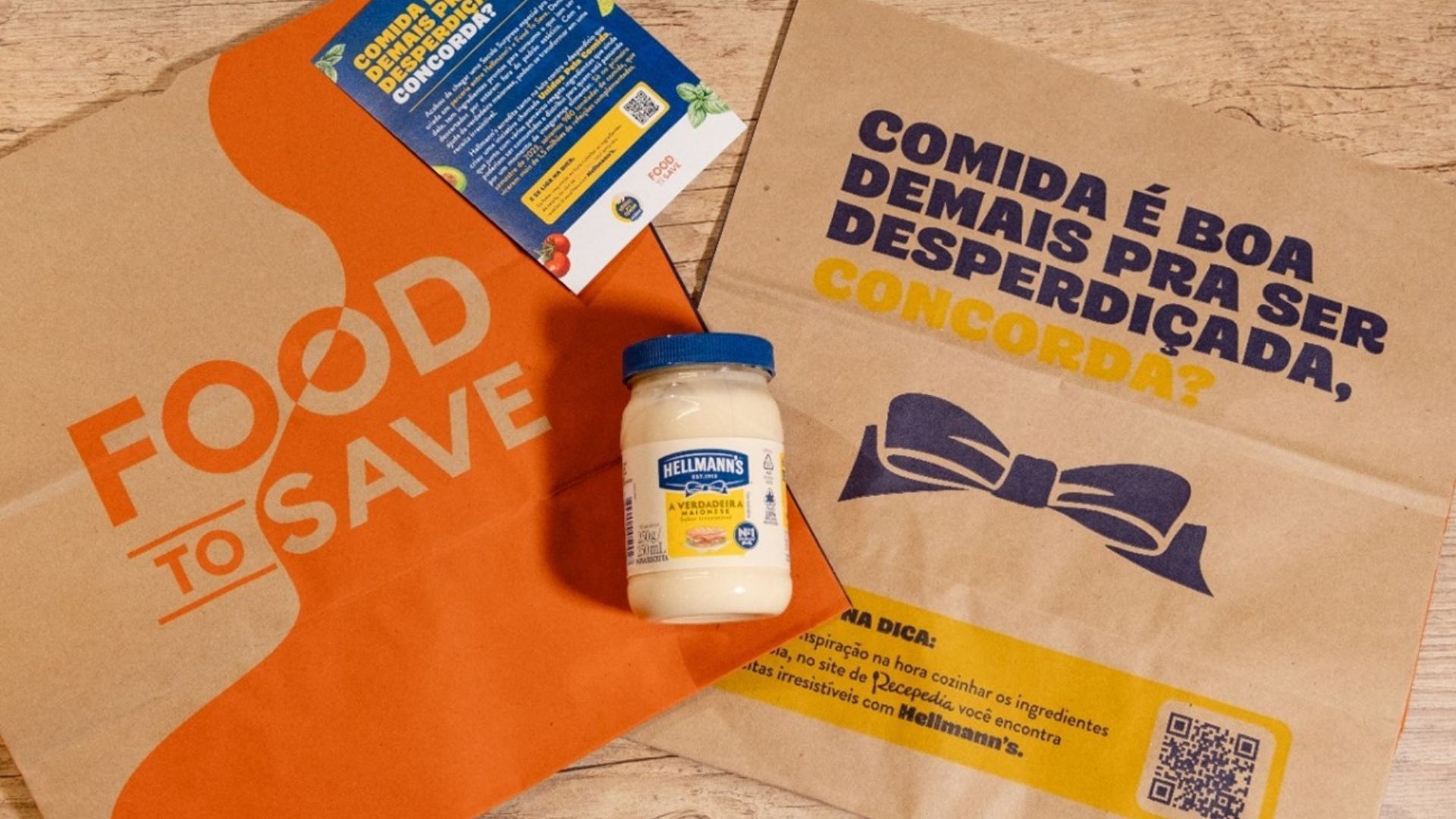Uma mesa de madeira clara comporta duas sacolas recicláveis: Uma sacola de papel kraft com o logo laranja escrito "Food To Save" , ao meio um pote de maionese Hellmann's com outra sacola em papel kraft com o logo de Hellmann's e a frase "Comida é boa demais para ser desperdiçada, concorda?"
