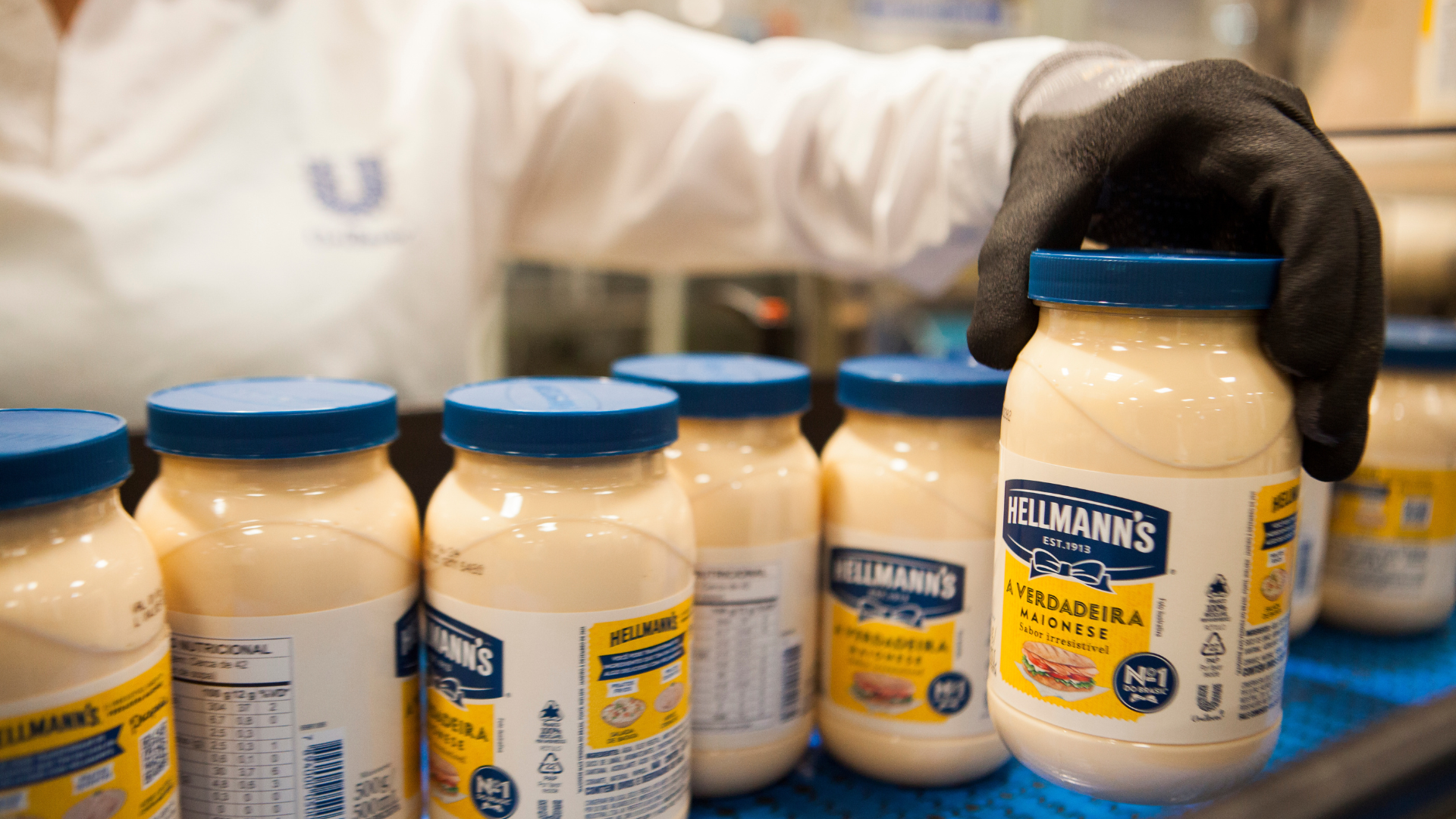 Várias embalagens de maionese Hellmann´s. Atrás, uma pessoa com roupa branca, da fábrica da Unilever, com o logo 