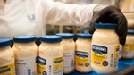 Várias embalagens de maionese Hellmann´s. Atrás, uma pessoa com roupa branca, da fábrica da Unilever, com o logo