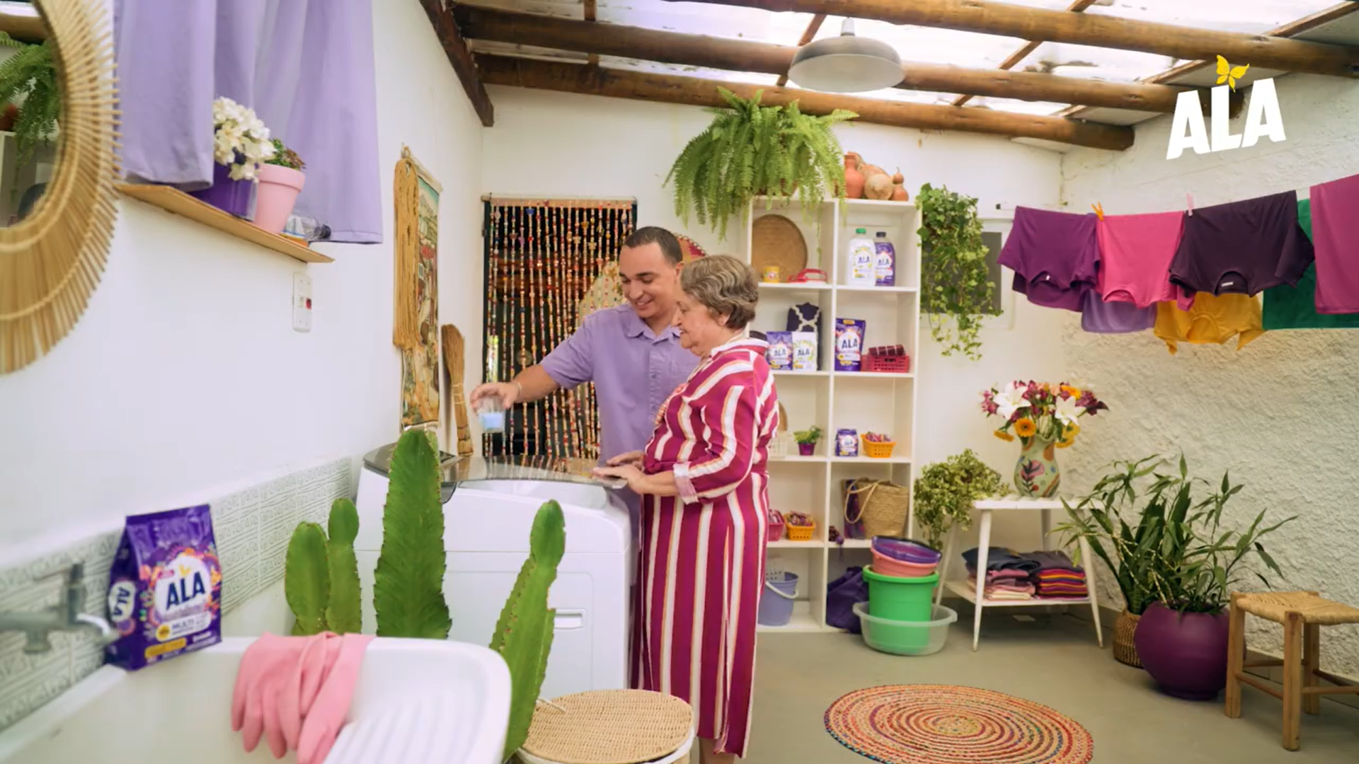 Uma lavanderia aconchegante, repleta de plantas, roupas coloridas penduradas e duas pessoas trabalhando juntas perto de uma máquina de lavar e detergente.