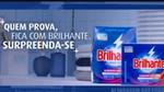 Duas embalagens do produto de limpeza Brilhante em destaque contra um fundo azul, com roupas dobradas e utensílios de limpeza.